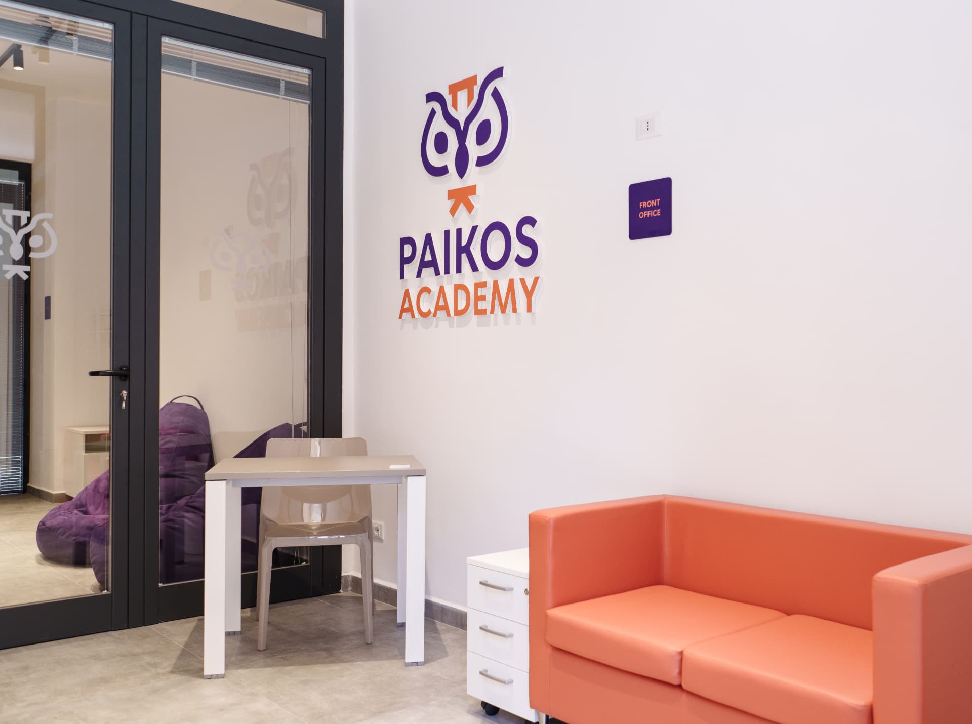 Front office di Paikos Academy con logo a parete, divano arancione e postazione accoglienza