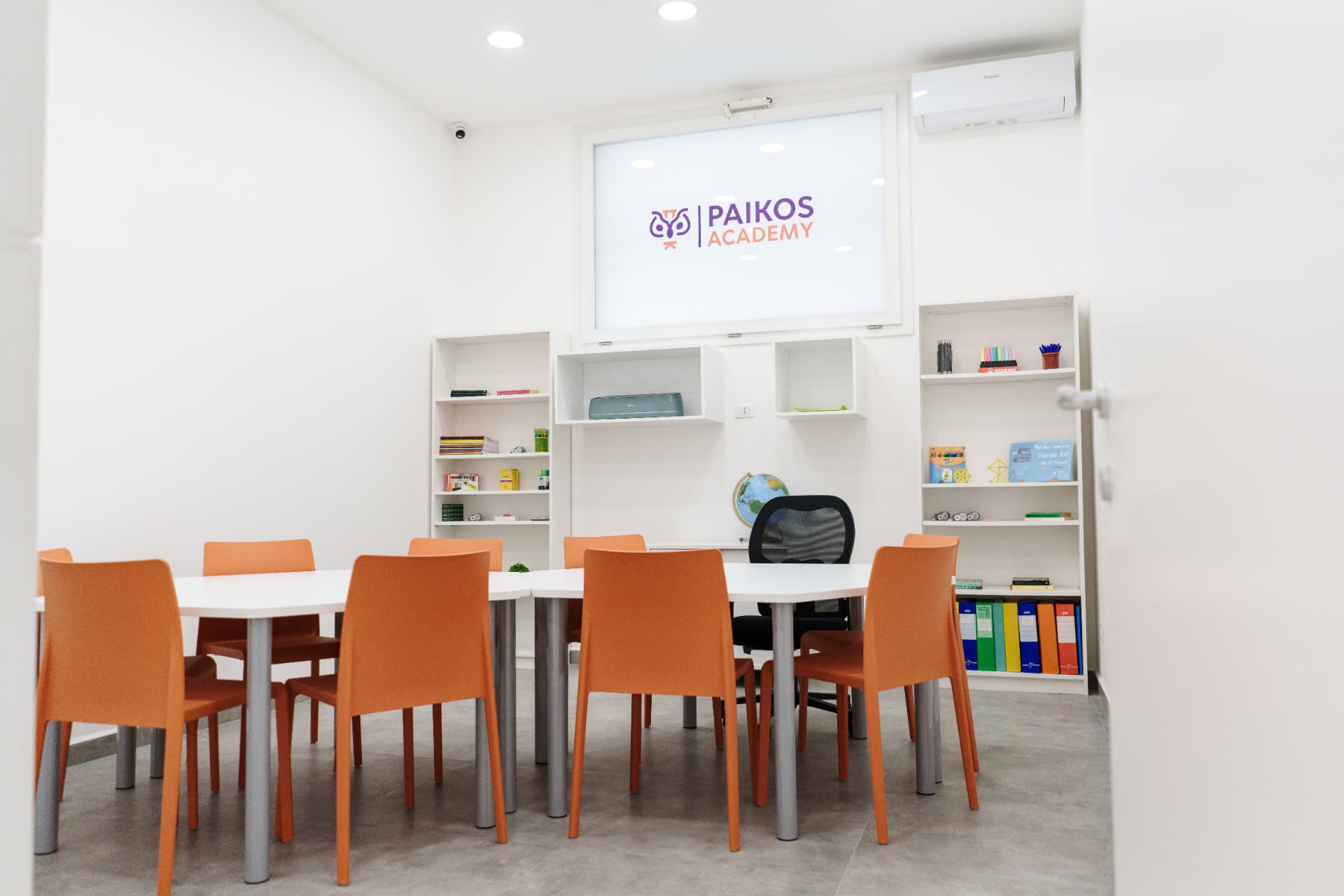 Aula Paikos Academy con logo sullo schermo, tavoli bianchi, sedie arancioni e libreria